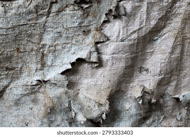 The surface texture of old, worn, and damaged plastic tarpaulin. The surface of the material shows signs of severe weathering, with the outer layer cracked, peeling, and discolored.