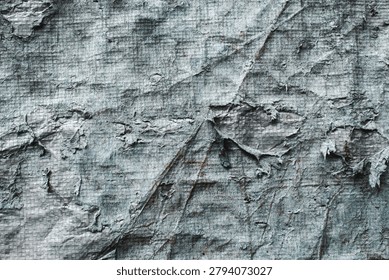 The surface texture of old, worn, and damaged plastic tarpaulin. The surface of the material shows signs of severe weathering, with the outer layer cracked, peeling, and discolored.