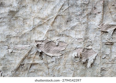 The surface texture of old, worn, and damaged plastic tarpaulin. The surface of the material shows signs of severe weathering, with the outer layer cracked, peeling, and discolored.
