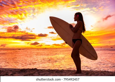 Surfer bikini girl on Hawaii beach holding surf board watching ocean waves at sunset. Silhouette of Asian sport woman over landscape, sky and clouds background. Summer vacation lifestyle.