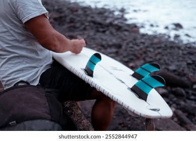 Surfer checking surfboard fins on a rocky beach, getting ready for surfing - Powered by Shutterstock