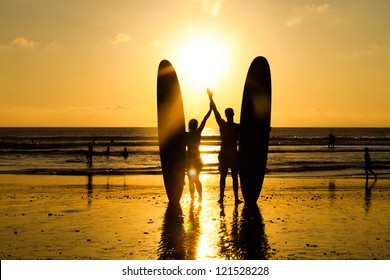 Surfer couple in silhouette holding long surf boards at sunset on tropical beach
