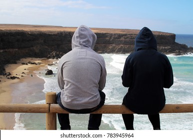 Surfer couple sitting on a fence wearing hoodies overlooking the waves hitting the beach and the cliffs