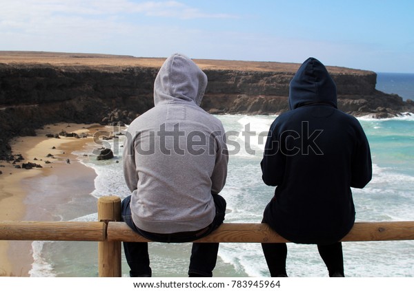 浜辺や崖を打つ波を見下ろすフードを着けたサーファー夫婦 の写真素材 今すぐ編集