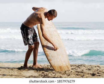 Surfer Dressed In Boardshorts Waxing Brown Surfboard On The Beach At Morning. Lifestyle.