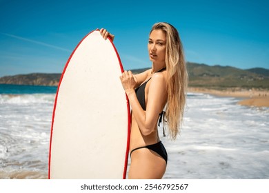 Surfer girl in black bikini holding surfboard while standing on scenic beach on sunny day. Attractive young female with long blond hair is enjoying summer vacation.  - Powered by Shutterstock