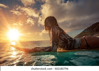A surfer girl watching sunset on a surboard floating in blue ocean near rocky shore