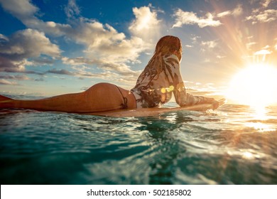A surfer girl watching sunset on a surboard floating in green blue ocean lit with sun flare