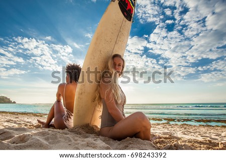 Surfer girls sitting back to back with surfboard in between in front of a breathtaking seascape on summer day, watching waves at beautiful Padang Padang beach, Bali, Indonesia