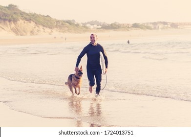 Surfer Having Fun With Best Friend German Shepherd Running And Playing On Dog-friendly Beach At Sunset. Summer Fun Surfing Vacation With Your Dog, Pet Friendly Trip And Outdoors Adventure Lifestyle.