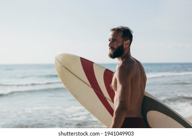 Surfer holding his surfboard on the beach - Hipster man standing on the beach and waiting big waves for surfing - Fit bearded man training with surfboard to sea - Lifestyle and freedom concept - Powered by Shutterstock