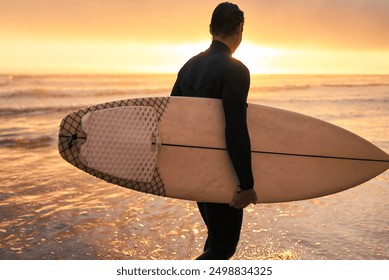 A surfer holding his surfboard stands at the shoreline, facing the ocean with the vibrant sunset in the background. - Powered by Shutterstock