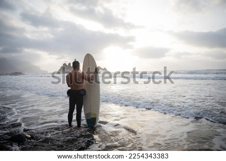 Surfer looks at the ocean while holding his surfboard at the beach, view from behind