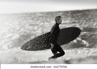 Surfer Silhouette Of A Man Holding His Surf Board, He Is Running On The Beach, He Is Wearing Wetsuit. Black And White Picture