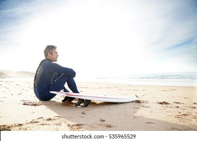Surfer sitting on sandy beach looking at the ocean