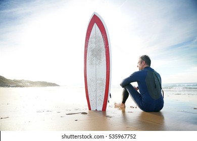 Surfer sitting on sandy beach, next to surfboard