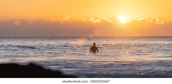 surfer sitting on their surfboard in the blue ocean and waiting for a wave at sunset in the evening watching the sun and clouds while the sky shines in orange. - Powered by Shutterstock