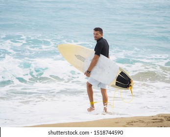 Surfer With A Surfboard Goes To The Ocean. Dressed In Shorts And Lycra. Beach On The Atlantic Ocean.