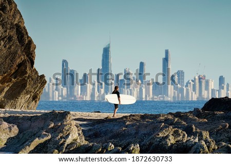 Surfer walking along a rocky coastline with the Gold Coast city skyline in the background.