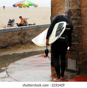 Surfer washes away sand under shower next to beach.