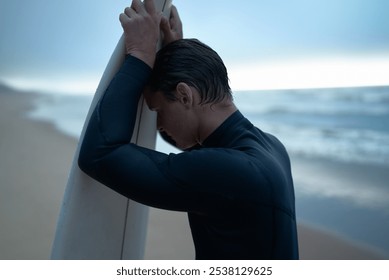 A surfer in a wetsuit leans on his surfboard, reflecting by the beach at dusk, conveying contemplation and solitude. - Powered by Shutterstock