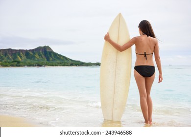 Surfer woman going surfing standing with surfboard on Waikiki Beach, Oahu, Hawaii. Female bikini girl walking with surfboard living healthy active lifestyle on Hawaiian beach. Asian Caucasian model.