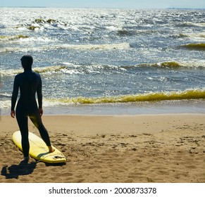 Surfer Woman With Surfboard Is Walking And Watching The Waves. Girl In Surfing Wet Suit Is Observing The Waves Of Cold Baltic Sea. Back View