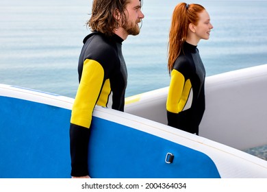 Surfers, Handsome Man And Redhead Woman Walking With Board On Beach. Water Sports. Caucasian Happy Couple Lead Healthy Active Lifestyle. Surfing. Summer Vacation. Extreme Sport. Portrait, Side View