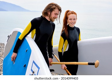 Surfers, Handsome Man And Redhead Woman Walking With Board On Beach. Water Sports. Caucasian Happy Couple Lead Healthy Active Lifestyle. Surfing. Summer Vacation. Extreme Sport. Portrait