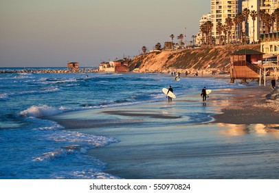 Surfers walking along the beach of Tel Aviv against the backdrop of a magnificent sunset and blue waves of the Mediterranean Sea.