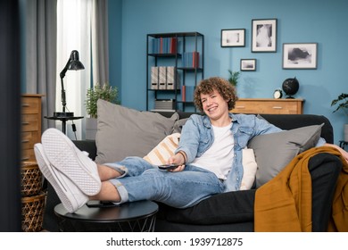 Surfing The Channels. Cheerful Young European Caucasian Teenager Male Holding Remote Control Watching Comedy On TV Laughing With Humorous Jokes While Sitting On The Couch At Living Room At Home