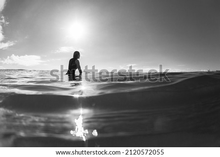 Surfing girl surfer silhouetted sitting on surfboard waiting in sunlight reflecting ocean water unrecognizable female rear behind  black and white photo  .