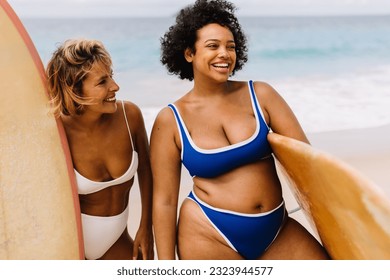 The surfing lifestyle comes to life as two women, surfboards in hand, stand together on the shore with happy smiles. Female surfers with diverse body sizes enjoy a fun beach experiences in the summer. - Powered by Shutterstock