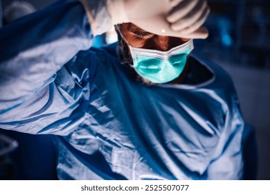 The surgeon in blue scrubs takes a contemplative moment to reposition his mask in a modern operating room, reflecting the careful and thoughtful nature of surgical procedures. Calm, focused. - Powered by Shutterstock