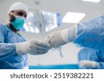 A surgeon in a blue surgical gown and gloves handing over a surgical instrument in a well-lit operating room, equipped with advanced surgical lights and equipment.