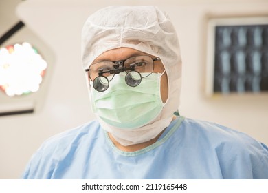A Surgeon In Blue Surgical Gown Suit Inside Modern Operating Room With Surgical Mask. Doctor Wear Loupe Magnified Glasses Prepare For Surgery In Private Clinic. Medical Concept.