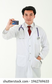 Surgeon Doctor Holding A Credit Card With A Smile On His Face, Expression Of Trust, Standing On White Background, Looking At The Camera. Healthcare, Health Insurance, Credit Card Service Concept.