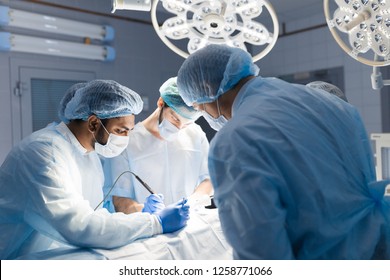 Surgeon Doctor Operating Using Electric Scalpel Wearing Blue Surgical Mask And Surgical Cap In Surgery Room With His Team Surgeons Operating