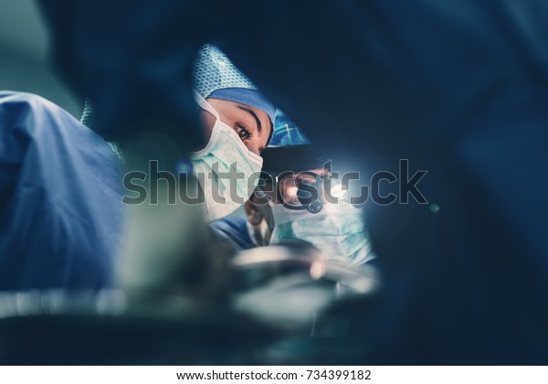 Surgeon doctor operating using special lamp\
lighting and glasses loupes wearing blue surgical mask and surgical\
cap in surgery room with his team surgeons operating live heart\
beating heart surgery