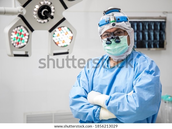 Surgeon doing crossed arm in blue surgical gown\
suit inside modern operating room with surgical mask. Doctor wear\
loupe glasses and head light prepare for surgery in private clinic.\
Medical concept.