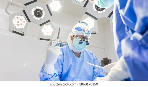 Surgeon Doing Surgery In Blue Surgical Gown Suit Inside Modern Operating Room With Surgical Mask. Doctor Wear Loupe Glasses And Head Light Suturing Inside Clinic With Scrub Nurse. Medical Concept.