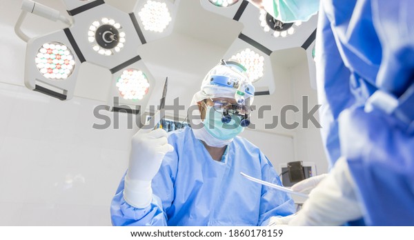 Surgeon doing surgery in blue surgical gown suit
inside modern operating room with surgical mask. Doctor wear loupe
glasses and head light suturing inside clinic with scrub nurse.
Medical concept.