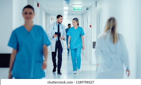 Surgeon And Female Doctor Walk Through Hospital Hallway, They Consult Digital Tablet Computer While Talking About Patient's Health. Modern Bright Hospital With Professional Staff.