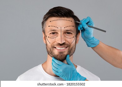Surgeon hands making marks on smiling man face. Aesthetic Cosmetology, Plastic Surgery concept. Plastic surgeon applying marks with pencil on handsome man face before surgery, grey studio background