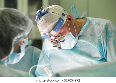 Surgeon and his assistant performing cosmetic surgery in hospital operating room. Surgeon in mask wearing loupes during medical procadure. Breast augmentation, enlargement, enhancement