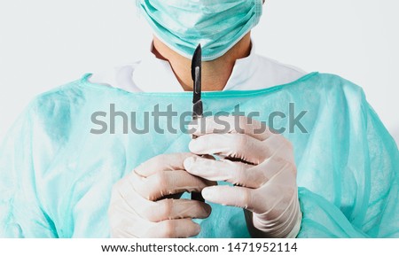 Surgeon holds scalpel dressed in a green surgical apron and mask on light background. Medical and pharmaceutical concept. Scalpel surgeon's scissors, just before surgery. Responsible work of surgeon.