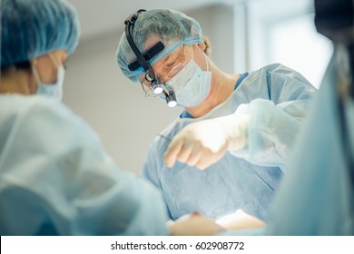 Surgeon performing cosmetic surgery on breasts in hospital operating room. Surgeon in mask wearing surgical loupes during medical procedure. Breast augmentation, enlargement, enhancement