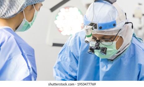 Surgeon Wearing Loupe Magnify Glasses Inside Modern Operating Room In Hospital.Doctor In Blue Surgical Gown Suit With Mask Protection.Medical Concept.Male Asian Doctor Perform Surgery.
