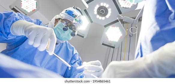 Surgeon Wearing Loupe Magnify Glasses Inside Modern Operating Room In Hospital.Doctor In Blue Surgical Gown Suit With Mask Protection.Medical Concept.Male Asian Doctor With Scrub Nurse Team.
