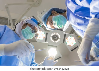 Surgeon Wearing Loupe Magnify Glasses Inside Modern Operating Room In Hospital.Doctor In Blue Surgical Gown Suit With Mask Protection.Medical Concept.Male Asian Doctor With Scrub Nurse Team.